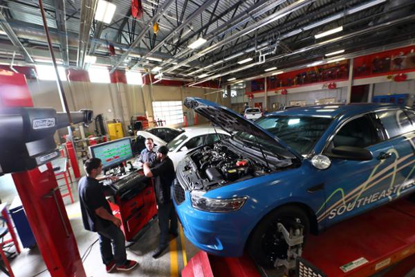 Three people in front of a computer monitor and a blue sedan with an open hood on a lift.