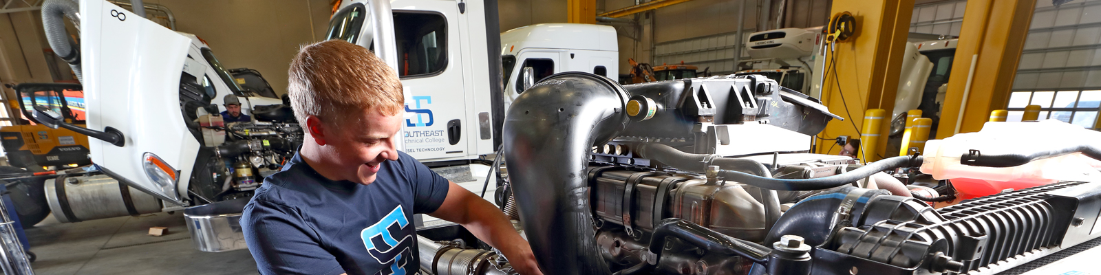 Student works on diesel engine with semi with open hood in background