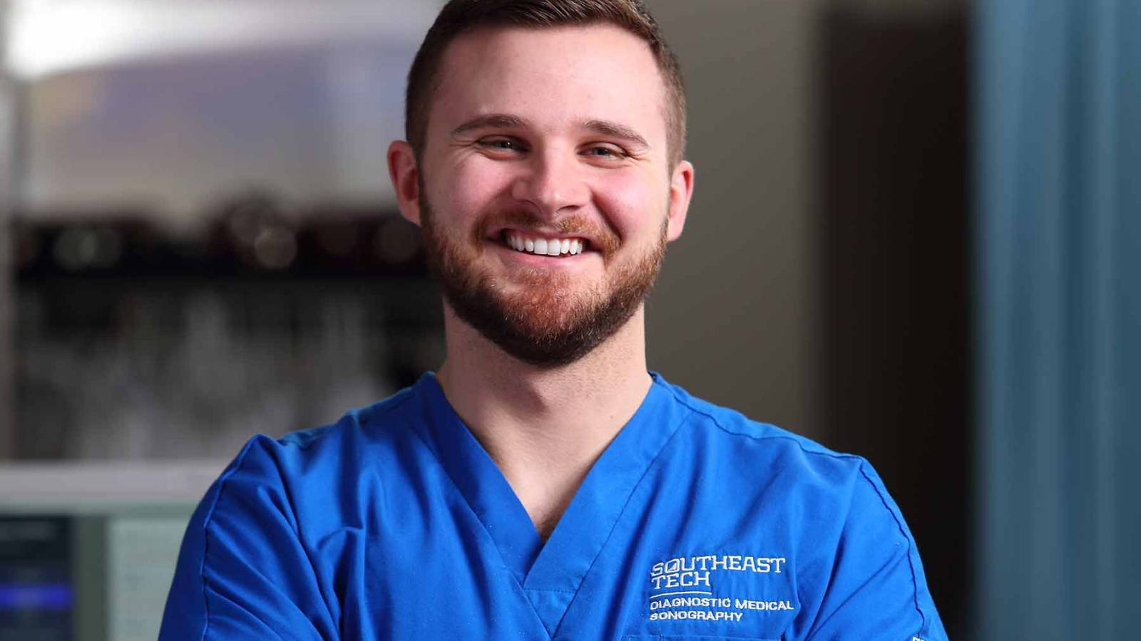 Male sonography student wearing blue scubs in exam room, smiling