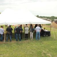 Group of people under a white tent on green grass with a construction site in the background. 