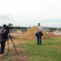 Camera and tripod interviewing student on dirt and green grass with large dirt pile and construction site in the background. 