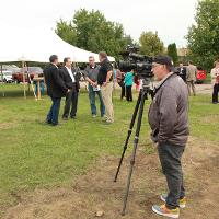 News reporter with camera and tripod, tent and people mingling in background. 